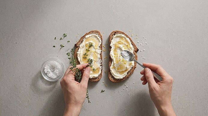 Zwei Hände bestreuen belegte Frischkäse-Brote mit Flockensalz von einem Löffel und frischen Thymianzweigen.