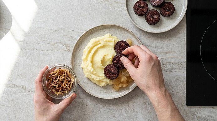 Zwei Hände legen gebratene Blutwurstscheiben auf einen Teller mit Kartoffelpüree und Apfelkompott; daneben ein Glas Röstzwiebeln und eine Schale mit weiteren Wurstscheiben.