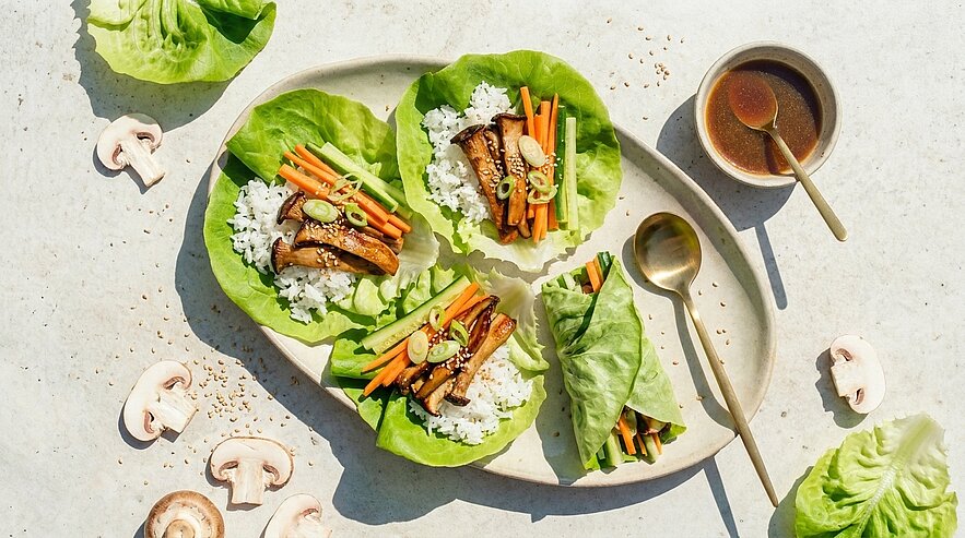 Drei Salatblätter als Wraps gefüllt mit Reis, gebratenen Champignons, Karottenstreifen, Frühlingszwiebeln und Sesam; daneben ein Schälchen Dip mit Löffel.