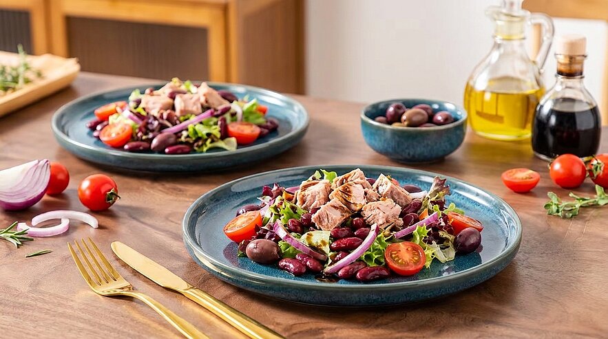 Zwei blaue Teller mit Thunfisch-Bohnensalat: gemischter Salat, Kidneybohnen, Cherrytomaten, Oliven und Thunfischstücken auf einem Holztisch.