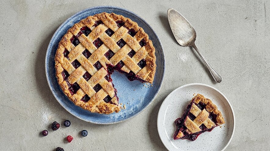 Gitter-Beerenkuchen auf blauem Teller, ein Stück herausgeschnitten und auf einem kleinen Teller serviert; daneben ein Löffel und einige verstreute Beeren.