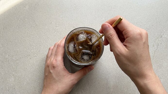 Zwei Hände halten ein Glas mit Eiskaffee und Eiswürfeln; die rechte Hand rührt mit einem Metallstrohhalm.