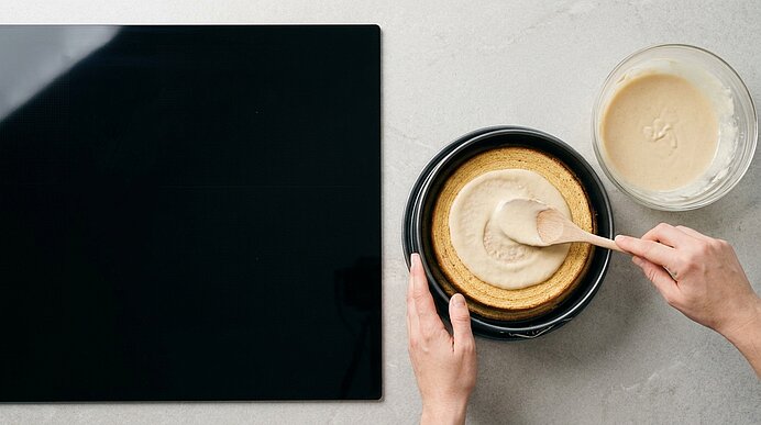 Hände verteilen eine helle Creme oder Teig mit einem Löffel gleichmäßig in einer runden Backform; daneben eine Schüssel mit restlichem Teig.
