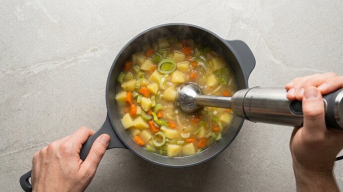 Draufsicht: Hände halten Topf und Stabmixer, mit dem eine Gemüsesuppe mit Kartoffel-, Karotten- und Lauchstücken püriert wird.