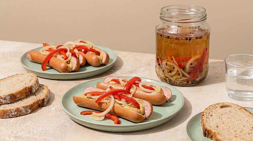 Drei Teller mit Würstchen in Brötchen, garniert mit dünnen Zwiebel- und roten Paprikastreifen; daneben ein Glas mit eingelegten Zwiebeln und Paprika sowie Brotscheiben.