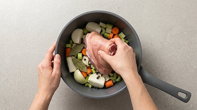 Zwei Hände legen ein rohes Fleischstück in einen Topf mit grob geschnittenem Suppengemüse (Karotten, Sellerie, Zwiebeln), Lorbeerblättern und ganzen Pfefferkörnern.