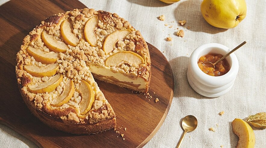 Ein frisch gebackener Kuchen mit einer goldenen Kruste, belegt mit Apfelscheiben und Streuseln. Der Kuchen ist teilweise angeschnitten und auf einem Holzbrett platziert. Daneben befindet sich ein kleines Gefäß mit Marmelade und ein Löffel.