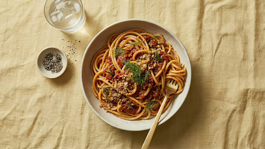 Eine Schüssel Spaghetti mit Tomatensauce, bestreut mit gehacktem Dill und Semmelbröseln, liegt auf einem hellen Tisch. Daneben befindet sich eine kleine Schale mit Pfeffer und ein Glas mit Eiswasser.
