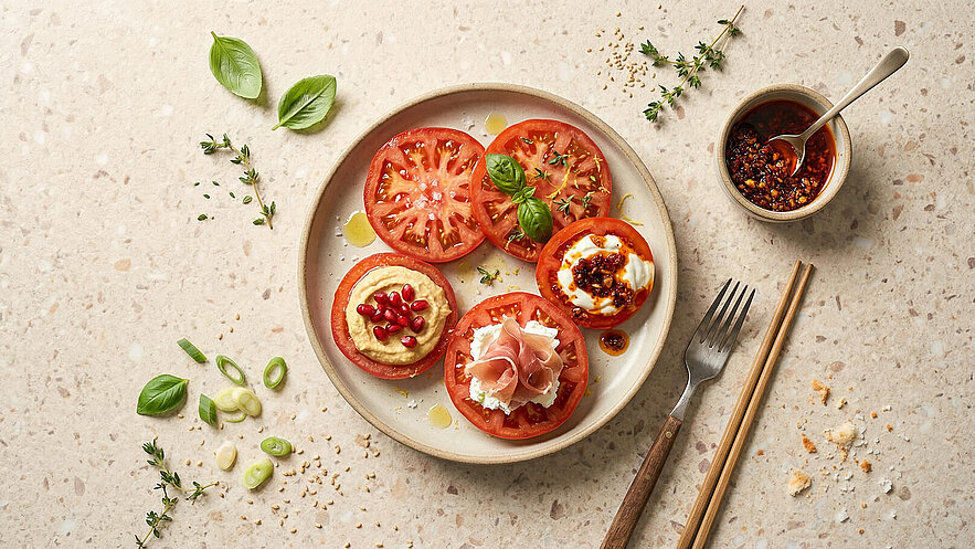 Ein Teller mit mehreren Scheiben Tomaten, darauf verschiedene Beläge wie Hummus mit Granatapfelkernen, Frischkäse und eine Sauce. Frische Kräuter wie Basilikum und Thymian sind dekorativ angeordnet. Neben dem Teller steht eine kleine Schüssel mit einer würzigen Sauce und Besteck.