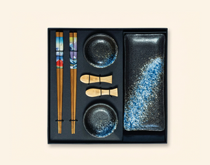 Sushi-Set in Geschenkbox: zwei Paar verzierte Essstäbchen, zwei runde Soßenschalen, zwei kleine hölzerne Fisch-Stäbchenhalter und eine längliche schwarze Platte mit blau-weißer Sprenkelung.