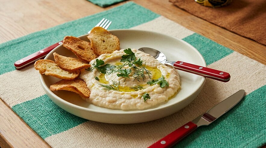 Schale mit cremigem Dip (Hummus-ähnlich), beträufelt mit Olivenöl und frischen Kräutern, serviert mit gerösteten Brotscheiben auf einem Teller; Besteck liegt daneben.