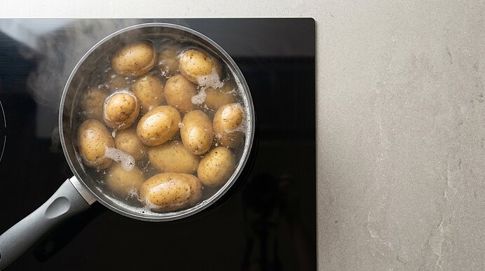 Draufsicht auf einen Topf mit kleinen Kartoffeln, die in kochendem Wasser auf dem Herd garen; sichtbarer Schaum und aufsteigender Dampf.