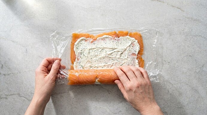 Zwei Hände rollen Lachs mit Dill-Frischkäse mithilfe von Frischhaltefolie auf einer hellen Arbeitsplatte auf.