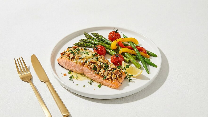 Ein Teller mit gebackenem Lachs, garniert mit einer knusprigen Kruste aus Mandeln und Kräutern. Dazu grüne Bohnen, gebratene Kirschtomaten und bunte Paprika. Ein Stück Zitrone liegt neben dem Lachs. Besteck aus Gold ist neben dem Teller platziert.