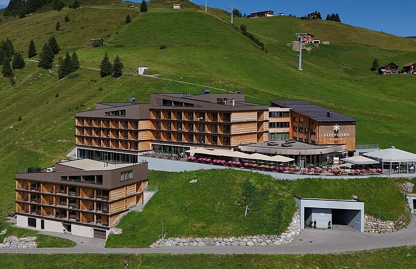 Das Bild zeigt ein modernes Hotel, das in einer bergigen Landschaft liegt. Umgeben von grünen Wiesen und Bäumen, hebt sich das Gebäude mit Holzverkleidung und großen Fenstern hervor. Vor dem Hotel sind Terrassen mit Sonnenschirmen und Blumenarrangements sichtbar.