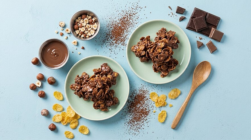 Auf einem blauen Hintergrund befinden sich zwei Teller mit schokoladigen Snacks aus Cornflakes und Nüssen. Neben den Tellern stehen eine Schüssel mit Nüssen, eine kleine Menge Schokolade, Kakaopulver und einige Cornflakes. Ein Holzlöffel liegt daneben.