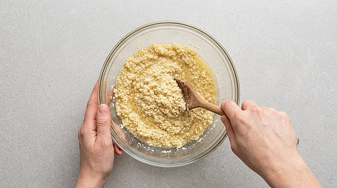 Zwei Hände rühren Riebelmais und Bouillon mit einem Holzlöffel in einer Glasschüssel auf grauer Arbeitsplatte.