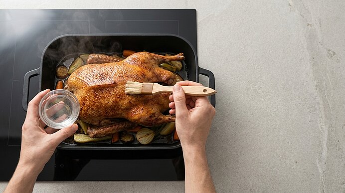 Zwei Hände bestreichen mit einem Pinsel ein frisch gebratenes, ganzes Hähnchen in einem Bräter mit geröstetem Gemüse.