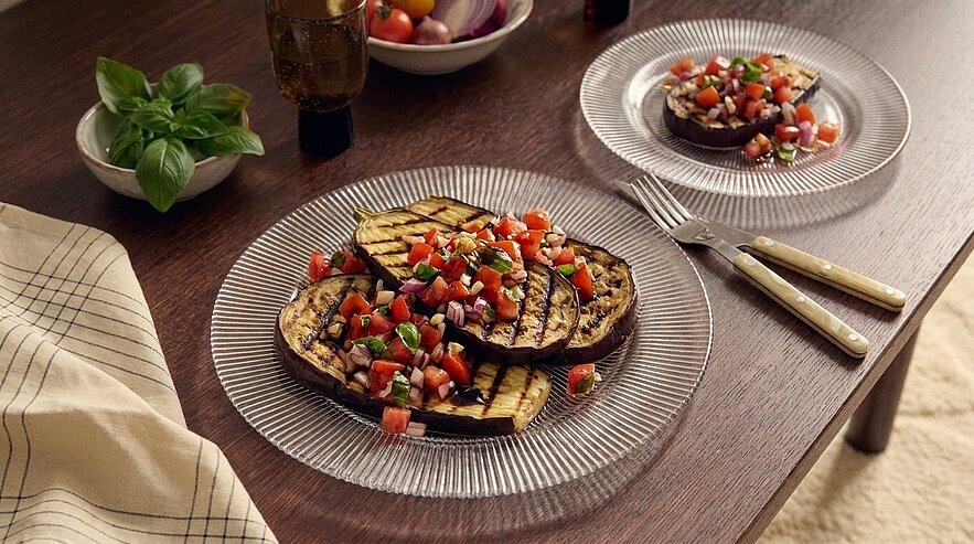 Auf einem Tisch liegen zwei Teller mit gegrillten Auberginenscheiben, die mit frischen Tomaten und Kräutern garniert sind. Neben den Tellern befindet sich ein kleiner grüner Basilikumbusch und eine Flasche Olivenöl.