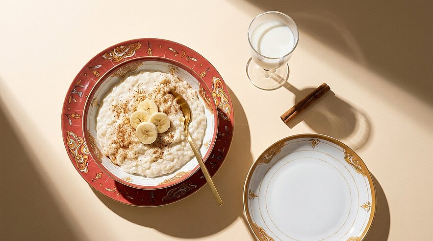 Schale Haferbrei mit Bananenscheiben, Zimt und Löffel; daneben Glas Milch und leerer Teller.