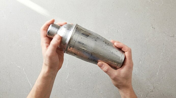 Zwei Hände halten einen silbernen, leicht abgenutzten Metall-Cocktailshaker über hellem Untergrund.