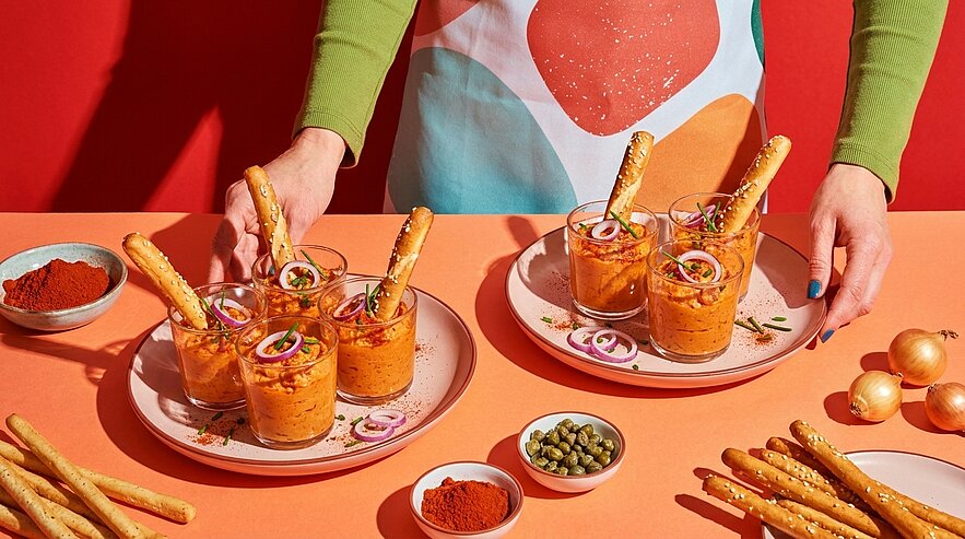 Sechs Gläser mit orangefarbenem Dip, jeweils mit Brotstange und roten Zwiebelringen garniert, auf zwei Tellern. Hände arrangieren die Teller auf einem Tisch mit kleinen Schälchen Gewürzen.