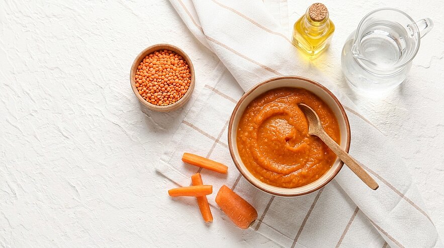 Schüssel mit orangefarbener Karotten‑Linsen‑Püree und Holzlöffel; daneben rohe Karottenstücke und eine kleine Schale mit roten Linsen.