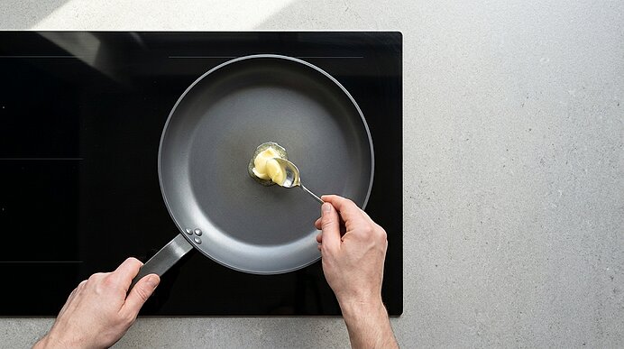 Blick von oben: Zwei Hände geben mit einem Löffel ein Stück Butter in eine antihaftbeschichtete Pfanne auf einem schwarzen Glaskeramik-Kochfeld.