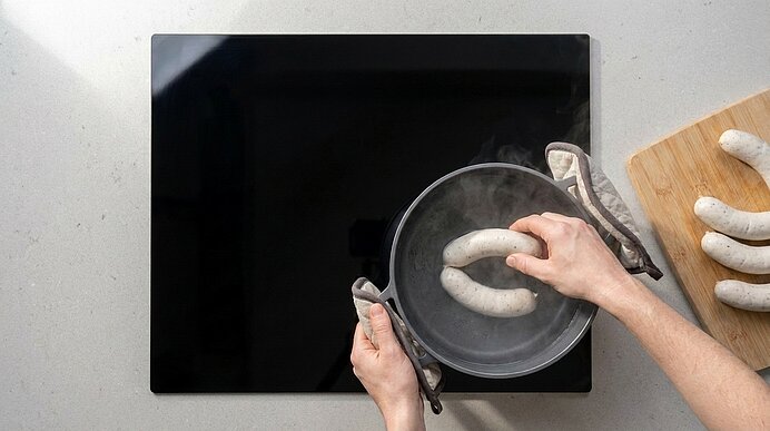 Zwei Hände legen eine weiße Wurst in einen dampfenden Topf mit heißem Wasser auf dem Herd; weitere weiße Würste liegen auf einem Schneidebrett.