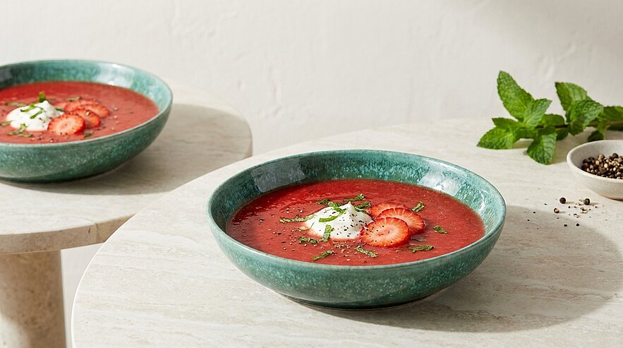 Zwei grüne Schalen mit kalter roter Erdbeer‑Gazpacho, im Vordergrund garniert mit einem Klecks Joghurt, frischen Erdbeerscheiben, Minzblatt und frisch gemahlenem schwarzem Pfeffer.