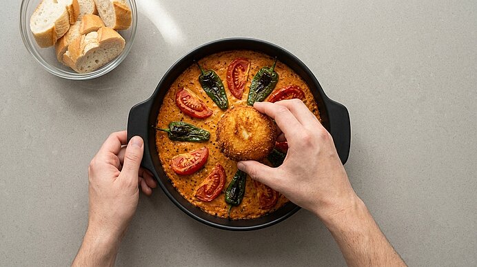 Eine Hand setzt die frittierte Burrata auf Pesto-Creme mit Tomaten und Paprika. Daneben liegen Baguettescheiben.