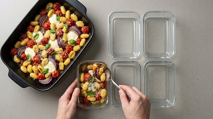 Hände füllen vier Glasbehälter mit gebackenem Gnocchi-Auflauf aus Kirschtomaten, roten Zwiebeln, Mozzarella, Basilikum und Parmesan aus einer ofenfesten Form.
