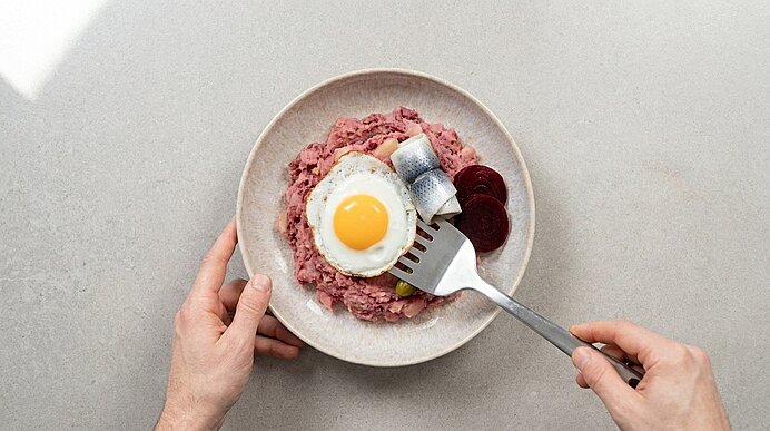 Teller mit rosa Fleisch‑Kartoffel‑Stampf (Labskaus), oben ein Spiegelei, rollierter Hering und Scheiben Roter Bete; zwei Hände halten Gabel und Teller.