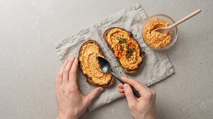 Zwei Hände bestreichen Roggensauerteigbrot mit Liptauer. Eine zweite Scheibe ist mit Schnittlauch und Paprika garniert.