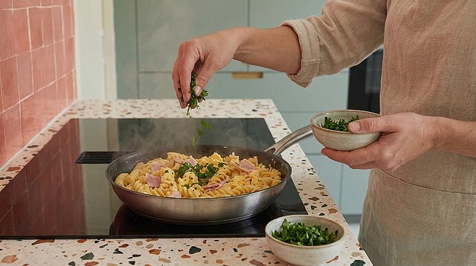 Hände streuen frisch gehackte Kräuter über Fusilli mit Schinken in einer Pfanne auf dem Herd.