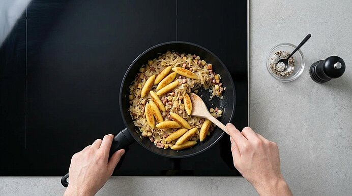 Zwei Hände mischen goldbraune Schupfnudeln mit einem Holzkochlöffel unter das Sauerkraut in einer mattschwarzen Pfanne.