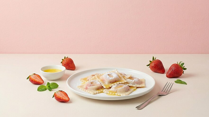 Ein Teller mit gefüllten Ravioli, leicht bestäubt mit Puderzucker, steht auf einem hellen Tisch. Neben dem Teller befinden sich frische Erdbeeren und eine kleine Schüssel mit einer gelben Sauce. Ein Besteckstück liegt bereit. Der Hintergrund ist in einem sanften Rosa.