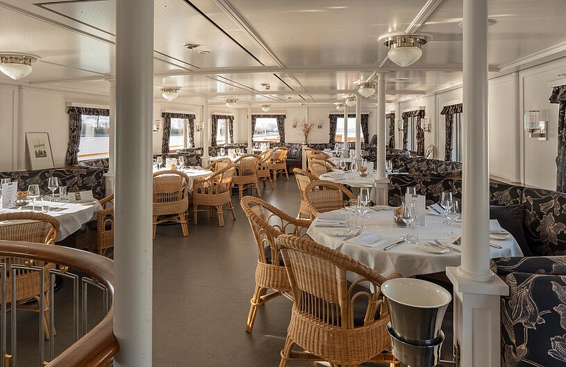 Ein elegantes Restaurant auf einem Schiff mit runden Tischen, eingedeckt mit Gläsern und Besteck. Die Wände sind in hellen Farben gehalten und kleine Fenster bieten einen Blick auf das Wasser. Korbsessel und stilvolle Vorhänge verleihen eine gemütliche Atmosphäre.