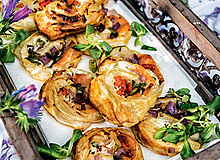 Ein rustikaler Holztablett enthält frisch gebackene, gefüllte Blätterteigrollen. Die Rollen sind mit Gemüse und Kräutern garniert und präsentieren sich appetitlich. Im Hintergrund sind dekorative Blumen sichtbar, die das Gericht umrahmen.