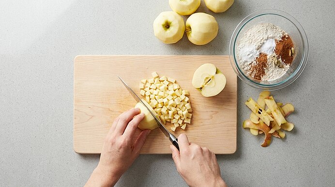 Zwei Hände würfeln einen geschälten Apfel auf einem Holzbrett; daneben liegen weitere geschälte Äpfel, Apfelschalen und eine Glasschüssel mit Mehl und Zimt — Vorbereitung zum Backen.