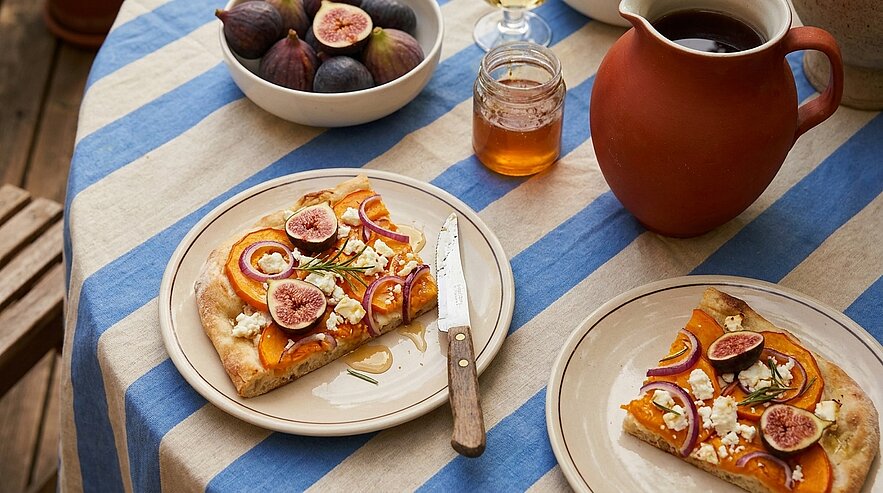 Zwei Teller mit Scheiben Flammkuchen belegt mit frischen Feigen, Käse und Kräutern auf blau-weiß gestreiftem Tisch, daneben Schale mit Feigen und Glas Honig.