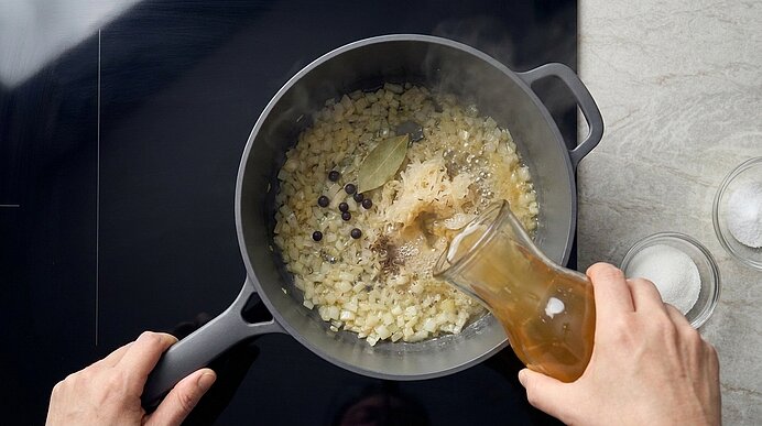 Top‑down-Ansicht: Zwei Hände gießen Brühe aus einer kleinen Kanne in einen schwarzen Topf mit angeschwitzten Zwiebeln, Lorbeerblatt und Wacholderbeeren; Dampf steigt auf.