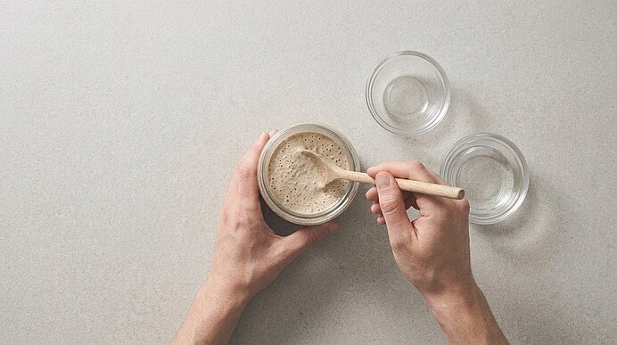 Zwei Hände verrühren mit einem Holzlöffel frischen Roggensauerteig-Starter mit Bläschen in einem Glas.