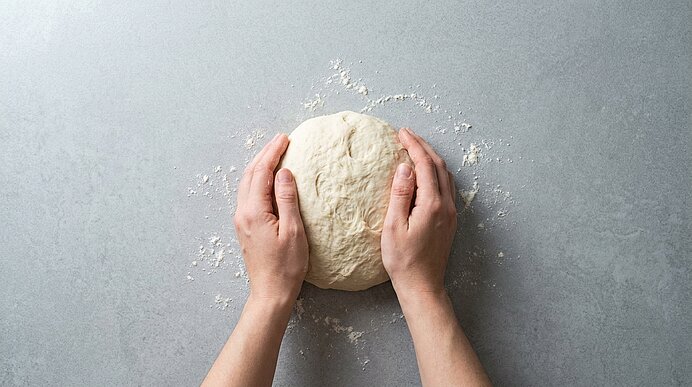 Zwei Hände kneten eine Kugel Hefeteig auf einer bemehlten, hellgrauen Arbeitsplatte.