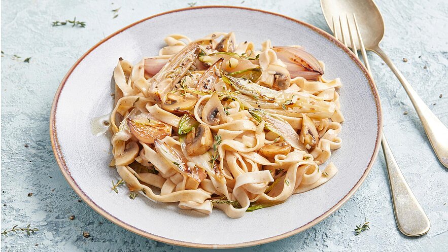 Ein Teller Pasta mit cremiger Soße, garniert mit karamellisierten Zwiebeln und Champignons. Die Nudeln sind dünn und leicht glänzend, umgeben von frischen Kräutern. Der Hintergrund ist hellblau, was den Fokus auf das Gericht lenkt.