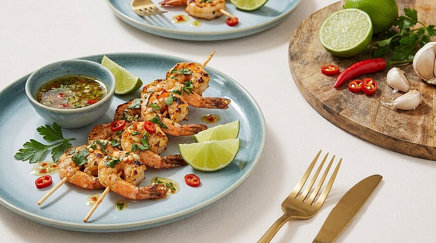 Auf dem Bild sind gegrillte Garnelen auf einem blauen Teller mit Limettenscheiben, frischen Kräutern und roten Chilischoten angerichtet. Neben den Garnelen befindet sich eine Schüssel mit Dip. Ein weiteres Gericht und eine Holzplatte mit Limetten und Knoblauch sind im Hintergrund sichtbar.