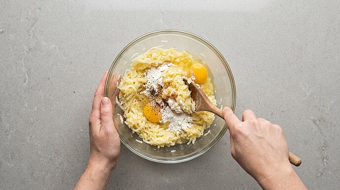 Zwei Hände rühren in einer Glasschüssel eine Mischung aus geriebenen Kartoffeln, Eiern und Mehl mit einem Holzlöffel.
