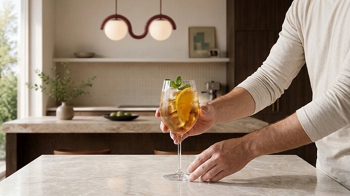 Oberkörper einer Person stellt ein Weinglas mit einem Aperitif, garniert mit Orangenscheibe und Minze, auf einer Küchenarbeitsplatte ab.