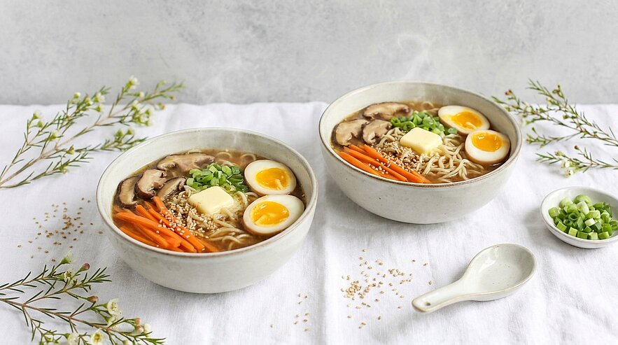 Zwei Schalen Ramen-Nudelsuppe stehen auf einem weißen Tisch. Die Suppen sind mit Stückchen von Karotten, Frühlingszwiebeln, Eiern und Butter dekoriert. Neben den Schalen liegt ein kleiner Löffel und eine Schale mit gehackten Frühlingszwiebeln. Dampf steigt aus den Schalen auf.
