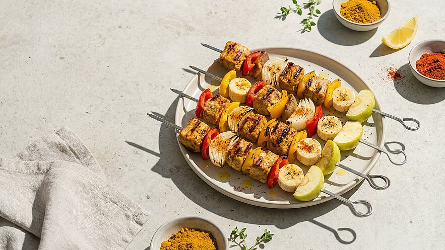 Ein Teller mit bunten Grillspießen, bestehend aus Fleisch, Paprika, Zwiebeln, Bananen und Äpfeln. Die Spieße sind appetitlich angerichtet und leicht angegrillt. Neben dem Teller liegen Gewürze und eine Zitronenscheibe.